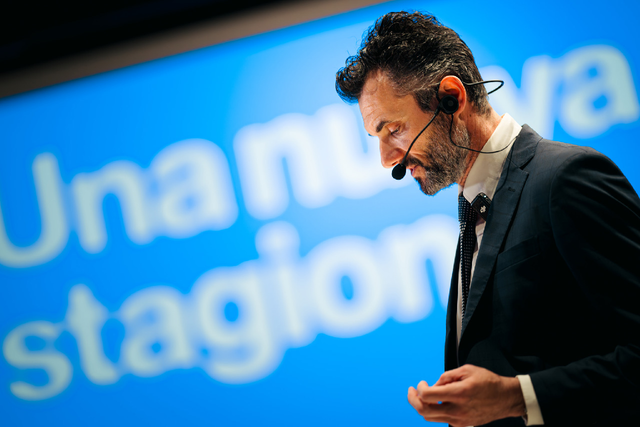 Primo piano di Matteo Biffoni durante il suo discorso sul palco. Sullo sfondo sfocato, la proiezione blu con il claim 'Una nuova stagione'.